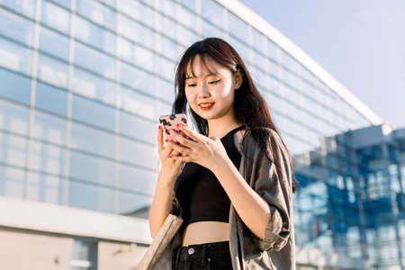 Close up lifestyle portrait of pleasant cute asian brunette girl with long dark hair, using her smartphone and looking for flight schedule, walking on the modern airport background.の写真素材