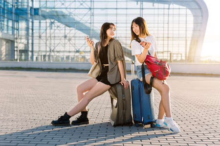 Summer portrait of two joyful asian women sitting on their luggage suitcases in front of airport building and using their phonesの写真素材
