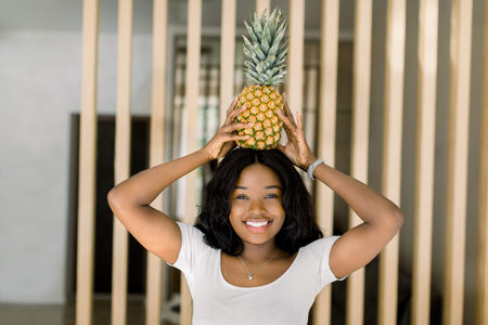 Portrait of joyful African girl in white t-shirt carries pineapple on head, looking at camera and...