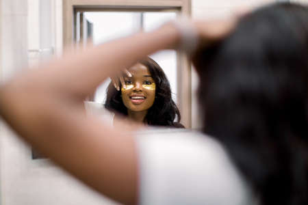Beauty treatment at home in the morning. African woman near the mirror doing beauty treatments on the face, putting golden eye patches on the area under the eyesの写真素材