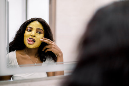 Portrait of young smiling African brunette woman in white t-shirt, standing in bathroom at home,...
