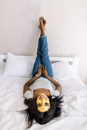 Young pretty African American woman with golden peel off facial mask resting, lying on bed, enjoying facial spa treatment at home. Skin care, beauty and spa at home.の写真素材