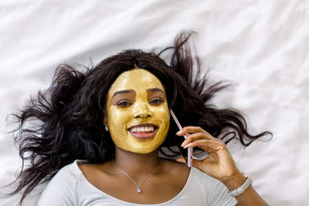 Top view of charming smiling African woman with golden clay facial mask lying on bed and using the cellphone, talking with her friend, while enjoying skin care treatment.の写真素材