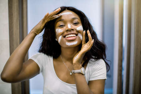 Pretty African woman looking in the mirror in bathroom, applying white mud facial mask, touching her face with hands and looking at camera. Beauty, skin care and treatment peeling.の写真素材