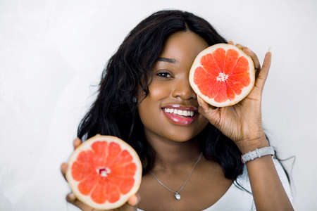 Close-up portrait of charming african girl holding halfs of grapefruit, hiding her eye. Amazing smiling dark skinned woman with grapefruit slices looking at camera on white background.の写真素材
