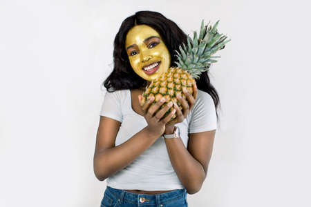 Portrait of cheerful young African American woman with golden cosmetic face mask, holding pineapple in her hands, isolated on white background. Skin care, fruit facial mask.の写真素材