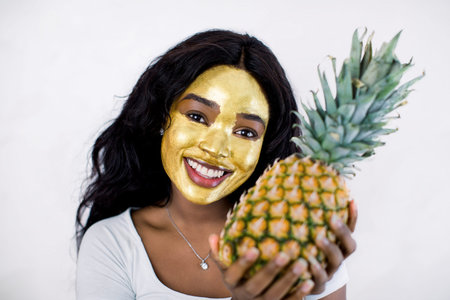 Close up portrait of beautiful sexy African girl with cosmetic peeling gold mask on her face, posing with fresh pineapple, on isolated white background.の写真素材