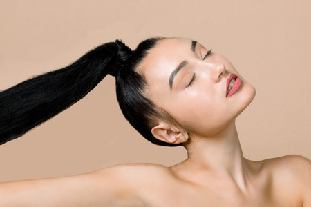 Young brunette Asian woman with beautiful black long hair in ponytail, posing on color studio beige...