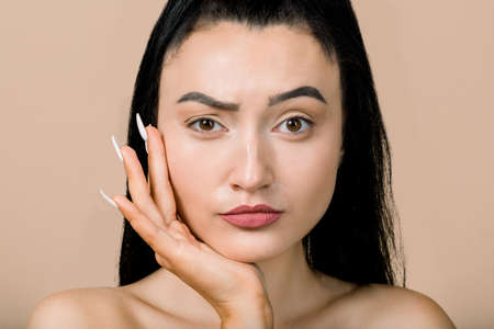 Asian beauty face. Closeup portrait of pretty young brunette woman, with confused and focused face, checking her skin and wrinkles. Skin care, acne and anti age treatment. Isolated on beigeの写真素材