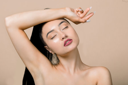 Face care, spa body treatment, cosmetology, beauty and healthy skin concept. Close up of asian relaxed naked woman, posing against beige background with eyes closed and one arm up, touching her head.の写真素材