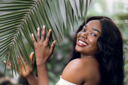 Natural beauty. Portrait of young lovely smiling African woman with long hair, posing to camera while standing near big exotic palm tree leaf in hothouse or garden.の写真素材