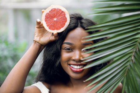 Relax, summer, holiday concept. Close up portrait of pretty smiling African girl, posing with grapefruit pie.ce and green palm tree leaf in greenhouse.の写真素材