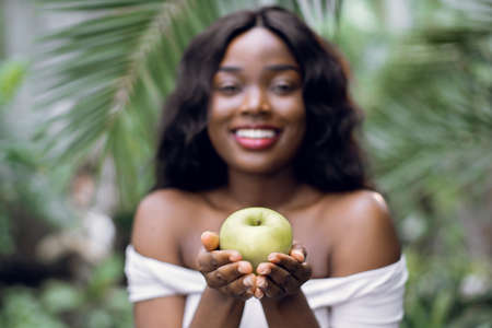 People, beauty, nature and healthy fruit. Blurred front close up view of young cheerful African woman in white t-shirt, posing with smile to camera, holding green apple, on exotic plants background.の写真素材