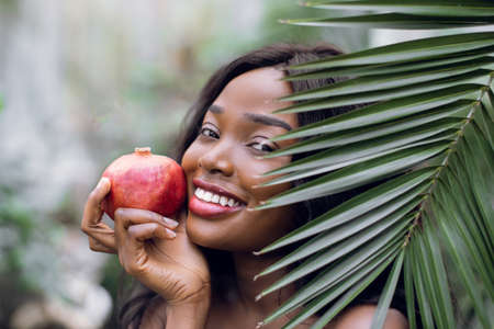 Close up portrait of young emotional beautiful afro american woman with pomegranate in her hands, looking out of green plam leaf indoors in greenhouse or garden. Summer and beauty concept.の写真素材