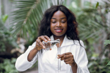 Young African American woman biologist in white coat pouring liquid from flask into test tube, while working in greenhouse with exotic plants and succulents. Focus on hands.の写真素材