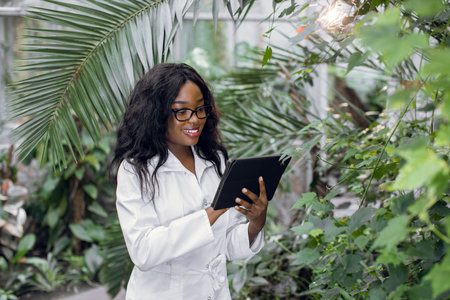Pretty smiling African American female agronomist supervising plants condition, working in greenhouse and making notes in clipboard.の写真素材