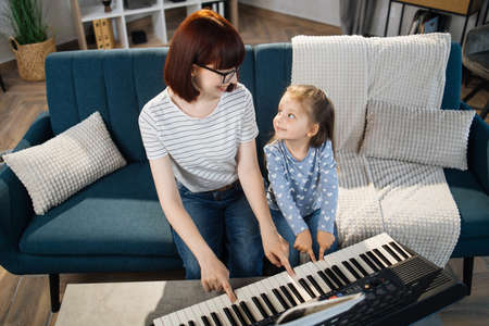 Little cute girl with music teacher having lesson at piano at school of music. Smiling young red hair woman looking at small kid girl sitting on the sofa and playing piano together in classroom.の写真素材