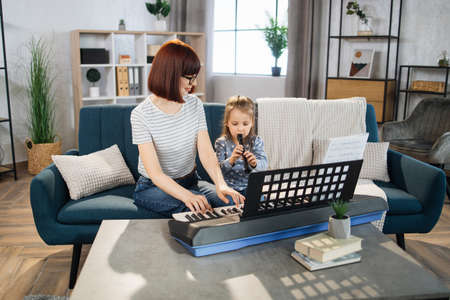 Beautiful young woman playing classic digital piano and her charming little daughter are smiling while playing flute at home. Mother teaching cute little musician girl to playing musical instruments.の写真素材