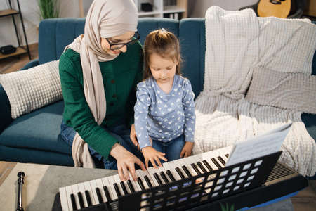 Little girl child learning to play classic digital piano with her young islam mom in hijab at home. Small kid preschool student playing musical instrument with muslim mother.の写真素材
