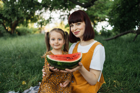 Summer time. Happy family of two, mother and her little girl at a picnic in the city park. Funny caucasian mom and daughter at the picnic eating watermelon.の写真素材