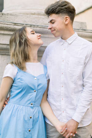 Portrait of smiling handsome man and pretty girl in blue dress standing on the background of ancient stone monument, holding hands and looking each other, outdoors in the city.の写真素材