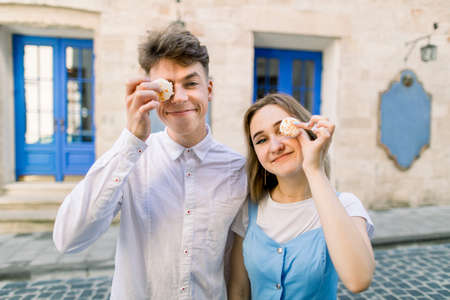 Horizontal shot of funny lovely young couple, walking outdoors in the city, posing to camera with tasty macarons, covering their eyes, on the background of vintage building with blue door.の写真素材