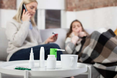 Close up of medicines, tablets, spray and drops for domestic treatment of flue or virus infection. Blurred view of woman calling to the doctor, checking her sick daughters temperature laying in bed.の写真素材