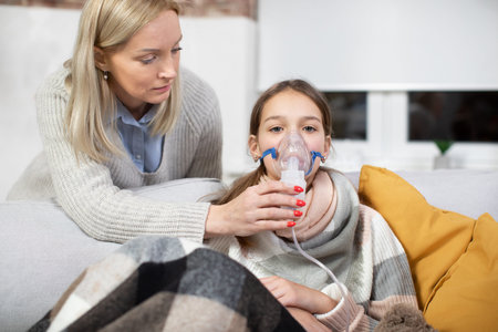 Mature Caucasian mom treats her teen daughter with nebulizer inhalation. Sick girl, suffering from...