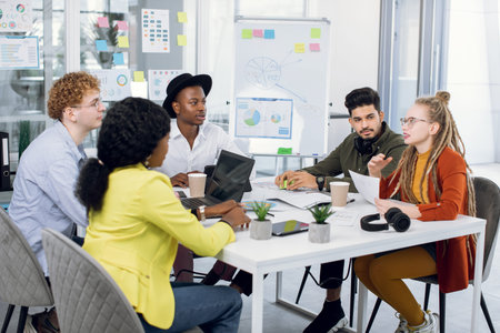 Group of five multiracial young people in casual outfit sitting at modern office and discussing new...
