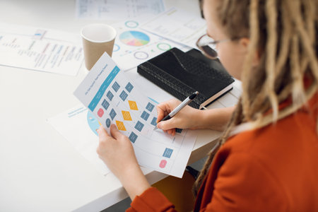 View from shoulder of young woman with dreadlocks working with data and chart at office. Hipster...