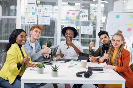 Smiling young freelancers in stylish outfit showing thumbs up on camera while sitting at office desk. Diverse creative team cooperating for doing successful startup.の写真素材