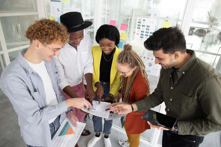 Close up top angle view of five multi ethnic students pointing with fingers on business plan that holding in hands. Group of creative people with gadgets start new common project.の写真素材