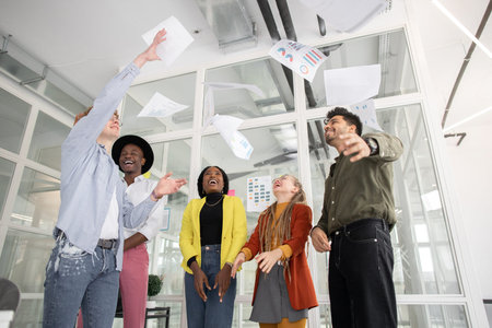Cheerful multiracial startup team of young colleagues throwing up documents at bright office....