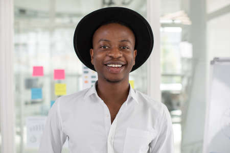 Close up portrait of positive handsome young african man wearing white shirt and black hat, standing at office in front of glass wall with stickersの写真素材