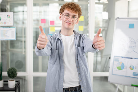 Positive young student or freelancer in glasses and casual wear showing thumbs up while standing at...