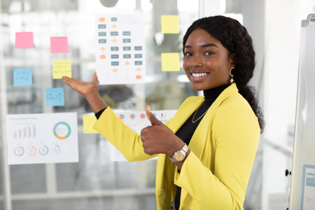 Close up of young attractive african woman in bright yellow jacket posing at office, showing thumb...