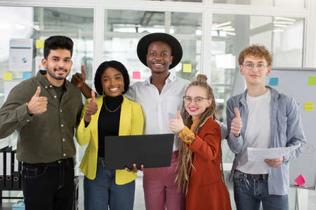 Group of five smiling multiethnic colleagues in stylish clothes showing thumbs up while standing together at bright office documents and modern laptop. Young diverse people celebrating success.の写真素材