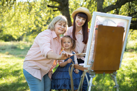Happy family three generations of women. Family painting outdoors. Cute little girl having fun with her mom and grandma, painting together a picture on a easel at spring garden.の写真素材