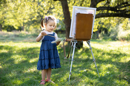 Painting on easel outdoors in the nature. Little cute talented excited kid girl artist, painting a picture on canvas, holding palette and brush and choosing a paint color.の写真素材