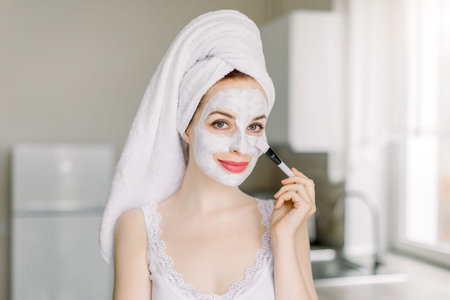 Pretty young Caucasian woman with hair wrapped in bath towel, posing to camera in modern home kitchen while applying white clay mud facial mask on her face with cosmetic brush. Spa and beauty at home.の写真素材