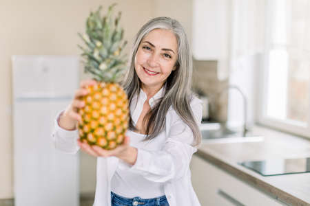 Healthy lifestyle, elderly people. Girl in the modern kitchen at home at her weekend time in the morning.Fresh pineapple in hands.の写真素材