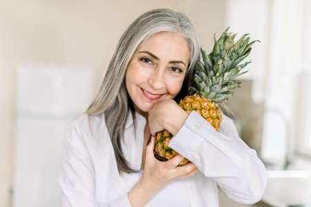 Close up horizontal beauty shot of happy senior gray haired woman holding fresh pineapple, looking at camera with smile, standing in modern light kitchen at home. People, portraits and healthy food.の写真素材