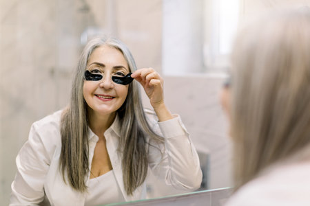 Facial mask and anti wrinkle therapy. Smiling elderly woman standing in front of bathroom mirror and...