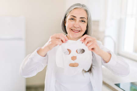 Face care and anti aging beauty treatments at home. Headshot portrait of elderly 60s woman with a cloth moisturizing mask, smiling to camera in light modern home interior.の写真素材