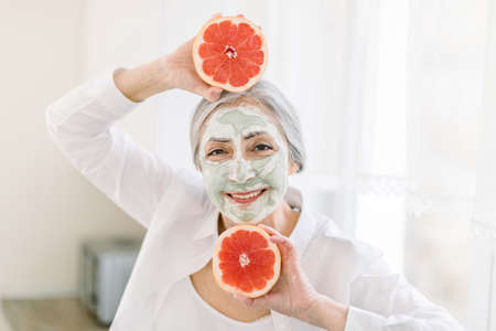 Close up beauty portrait of pretty elderly grai haired woman with mud clay facial mask, holding near face two fresh grapefruit slices, smiling to camera. Promo of facial masks and skin care.の写真素材