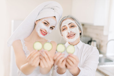 Morning skin care routine, family spa day. Pretty senior gray haired woman and her young attractive granddaughter making clay facial masks and posing to camera in kitchen with cucumber slices.の写真素材