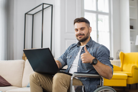 Handsome disabled man sitting in wheelchair with laptop on knees and looking at camera. Caucasian...