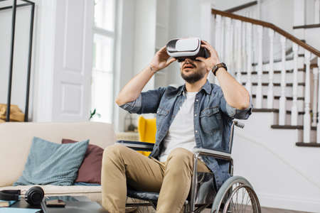Disabled man watching virtual video using goggles of augmented reality while sitting in wheelchair at home. Concept of full technology opportunities for people with special needs.の写真素材
