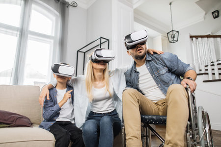 Attractive mother hugging cute son and bearded father in wheelchair while using vr headset at cozy home. Concept of happy family, modern technology and leisure time.の写真素材