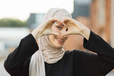 Smiling arabian muslim woman in hijab and black clothes posing on urban background. People religious Islam lifestyle concept. Showing shape heart with hands, heart-shape sign.の写真素材